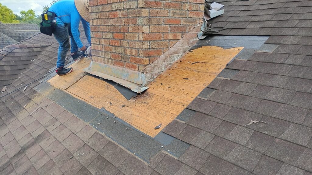 A worker in blue attire focuses on repairing a roof around a brick chimney, carefully addressing the Houston roofing needs. Old shingles have been removed, revealing plywood beneath. The scene unfolds against brown roof shingles, embodying essential roofing maintenance.