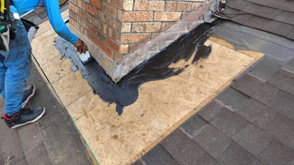 A worker in blue attire is applying black sealant around the base of a brick chimney on a roof, ensuring waterproofing for Houston roofing. The roof surface features a plywood section and brown shingles, and the worker uses a tool to expertly spread the sealant.