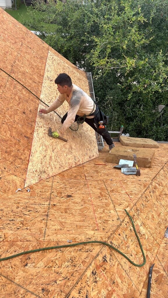 A person in a harness diligently works on a pitched wooden roof, installing boards with precision, reminiscent of Houston roofing experts. Nearby, tools like a drill and hammer lie ready amidst the backdrop of lush green trees.