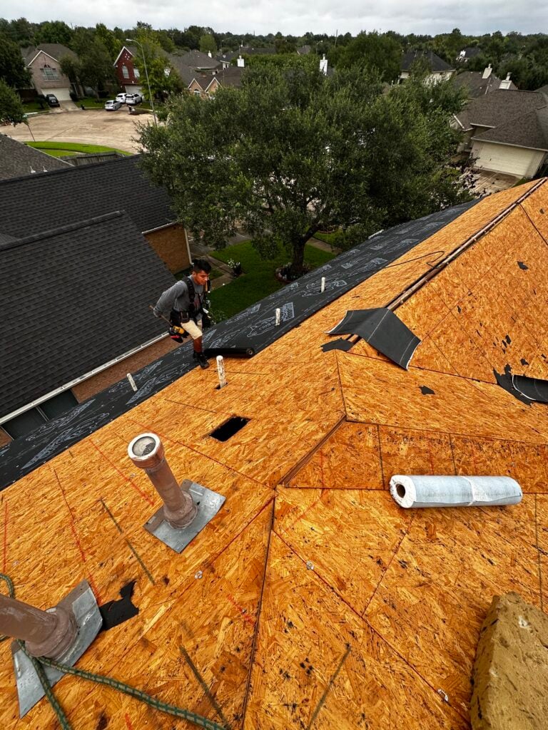 A worker in a gray jacket and safety harness is expertly installing underlayment on a wooden roof. Surrounded by vent pipes and roofing materials, this Houston roofing project overlooks suburban houses and trees in the distance.