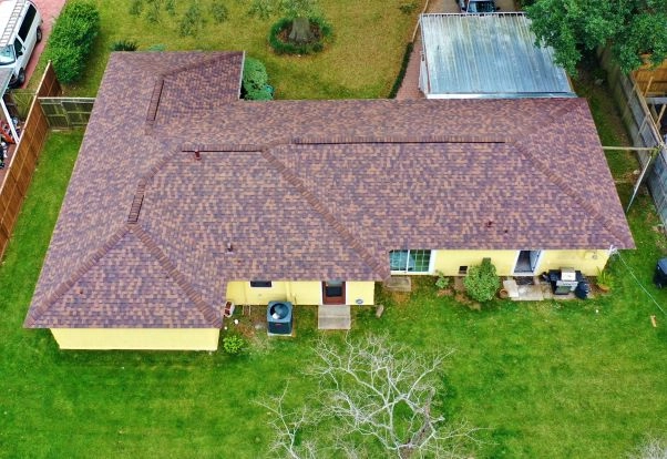 Aerial view of a single-story yellow house with brown shingled roof, surrounded by a grassy yard. The backyard has a tree with bare branches, a small patio, a central HVAC unit, and various small plants near the house. A metal-roofed structure is adjacent.