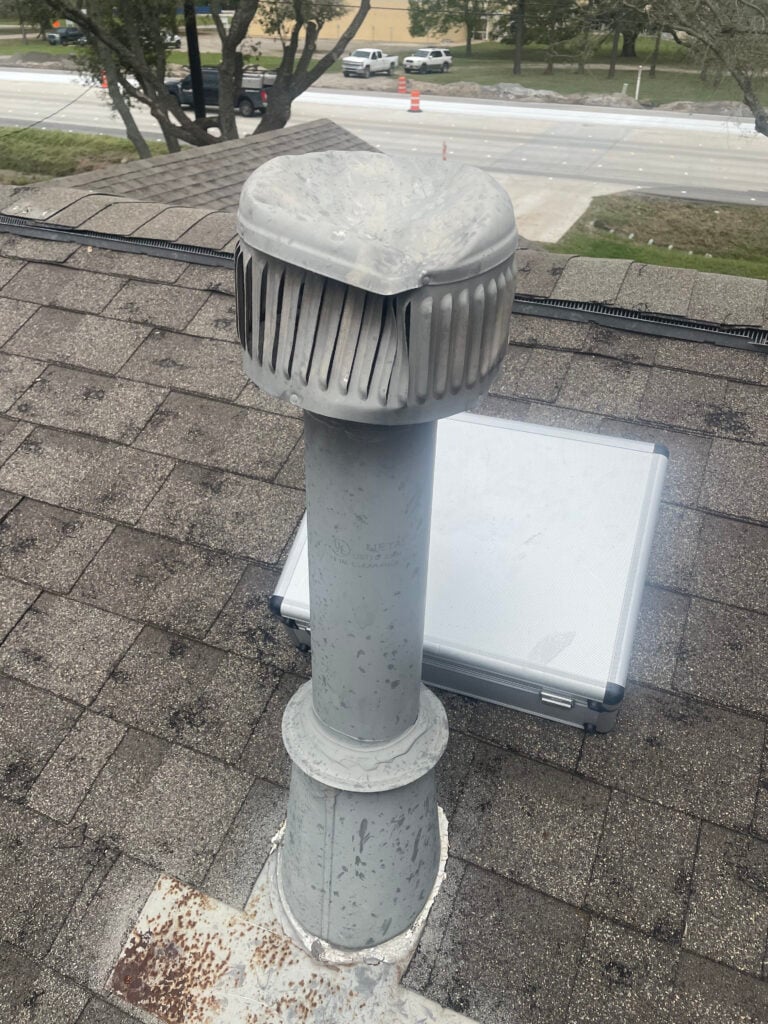 A weathered metal exhaust vent, with a cylindrical body and ridged cap, sits atop a shingled rooftop in Houston. Rust adds character to its surface. A solar panel is partially visible in the background, alongside a road and trees.