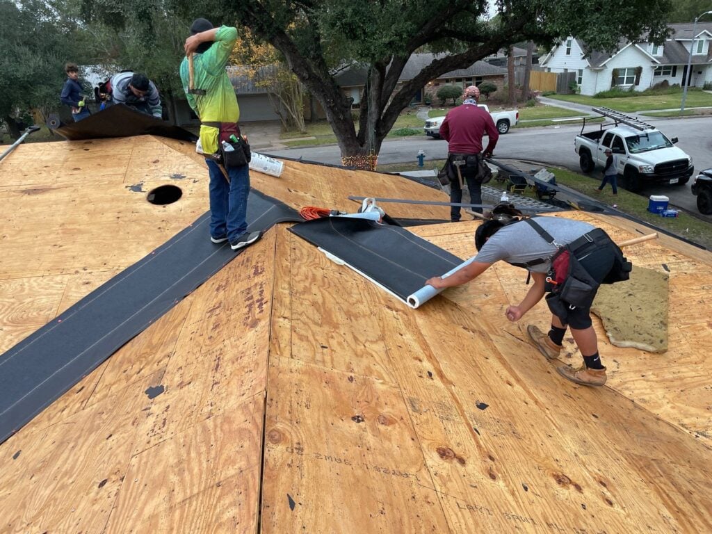 Workers applying roofing underlayment on a wooden roof in Houston. One person spreads the material while others prepare the area, surrounded by trees and parked vehicles on a residential street.