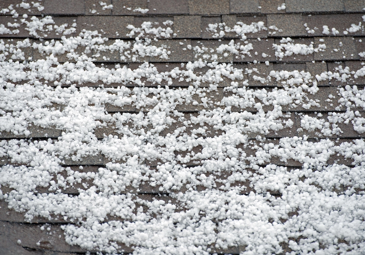Close-up of a roof covered with numerous small hailstones scattered across the shingles. The hailstones vary in size and are spread unevenly, creating a textured appearance against the dark background of the Houston roofing material.