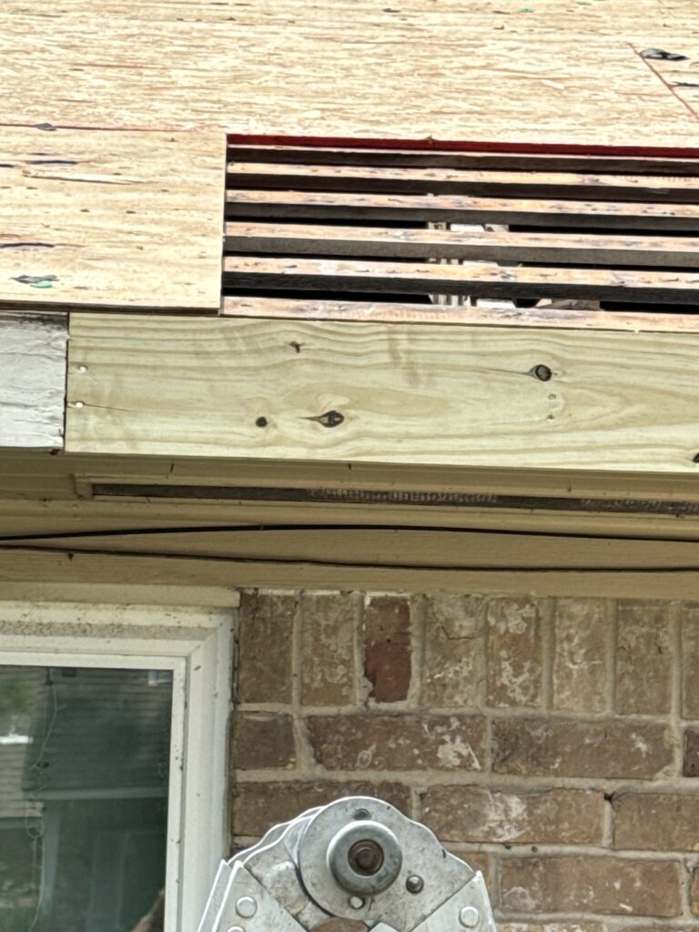 A brick house in Pecan Grove showcases a corner with new plywood roofing being installed as part of roof repairs. Partially completed siding and shingles are visible, with a white window on the left side. A metal tool or fixture is in the foreground, highlighting ongoing work.