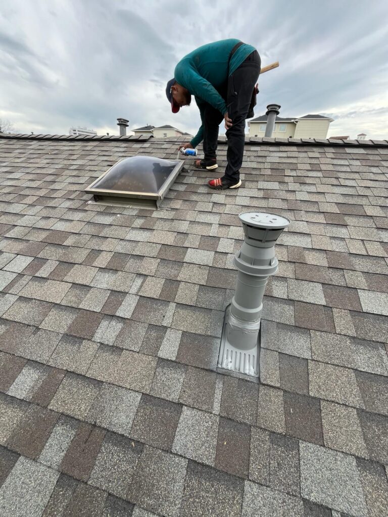 A Houston roofer in a teal sweater and cap is expertly applying sealant around a skylight on the shingled roof. A vent pipe stands nearby under the overcast sky.