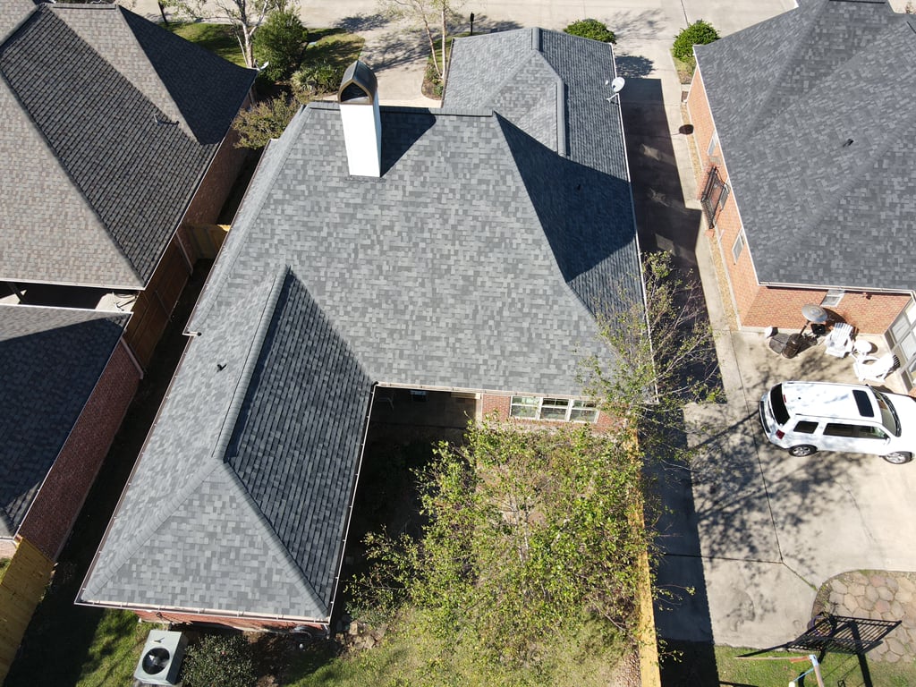 Aerial view of a suburban home with a gray shingle roof, hinting at recent roof replacement. The house, complete with a white chimney, sits among similar homes. A driveway houses a parked white car, while trees and a small garden enhance the four corners of the surrounding area.
