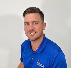 A man with short dark hair and a beard is posing against a plain white background. He is smiling and wearing a blue polo shirt with a logo on the chest that reads