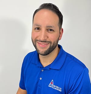 A man with short dark hair and a trimmed beard is smiling while wearing a blue polo shirt with a company logo that reads
