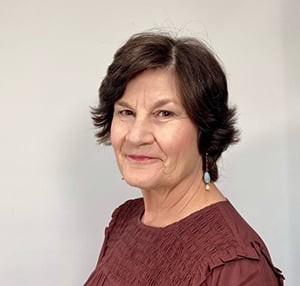 A middle-aged woman with short, dark hair wearing a maroon top stands against a plain background. She has light makeup and is smiling slightly. She is wearing dangling turquoise earrings.