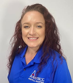 A woman with long, curly reddish-brown hair is smiling at the camera. She is wearing a blue collared shirt with the logo and text