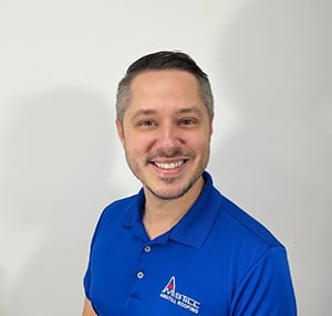 A man with short hair and a neatly trimmed beard is smiling at the camera. He is wearing a blue polo shirt with a logo that reads