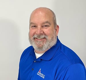 A smiling man with a bald head and a full gray beard is wearing a bright blue polo shirt with a logo on it. The background is plain and white.