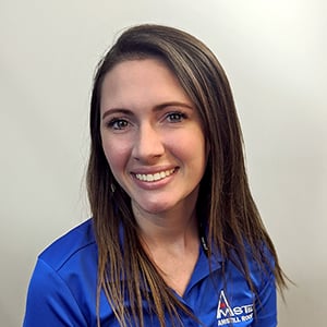 A woman with long, straight brown hair is smiling at the camera. She is wearing a blue collared shirt with a logo on it. The background is plain and light-colored, likely an office setting.