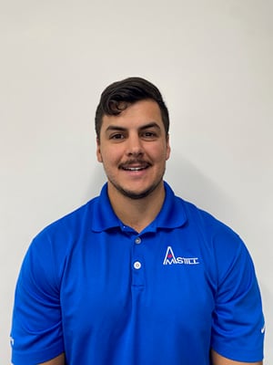A man with short dark hair and a mustache is wearing a bright blue polo shirt with a logo on it. He is smiling and standing in front of a plain white background.