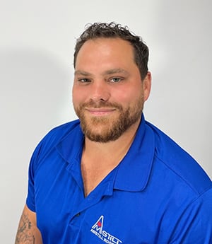 A person with short curly hair and a beard is smiling at the camera. They are wearing a blue collared shirt with a logo that includes the text