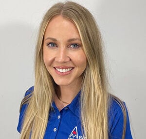 A woman with long blonde hair is smiling at the camera. She is wearing a blue polo shirt embroidered with a logo that includes the letters