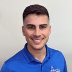 A man with short dark hair and light skin smiles at the camera. He is wearing a bright blue polo shirt with a logo on the left chest that appears to be related to steel or construction. The background is plain and light-colored.