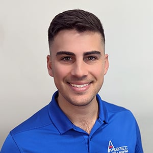 A man with short dark hair and light skin smiles at the camera. He is wearing a bright blue polo shirt with a logo on the left chest that appears to be related to steel or construction. The background is plain and light-colored.