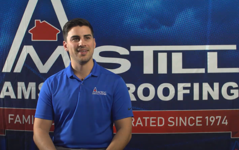 A man wearing a blue polo shirt with the "Amstill Roofing" logo sits and smiles in front of a large "Amstill Roofing" backdrop. The backdrop also displays the text "Family Owned and Operated Since 1974.