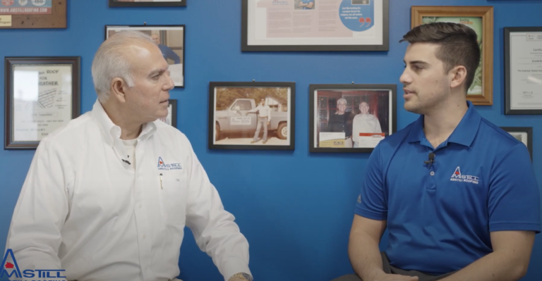 Two men, one older and one younger, are seated and engaged in conversation against a blue wall adorned with various framed certificates, photos, and documents. Both wear shirts with the Mistill company logo. The older man is wearing white, and the younger man is in blue.