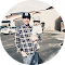 A person wearing a cap and a plaid shirt is holding a small child dressed in a white outfit. They are standing in a paved area with buildings and cars in the background on a sunny day.