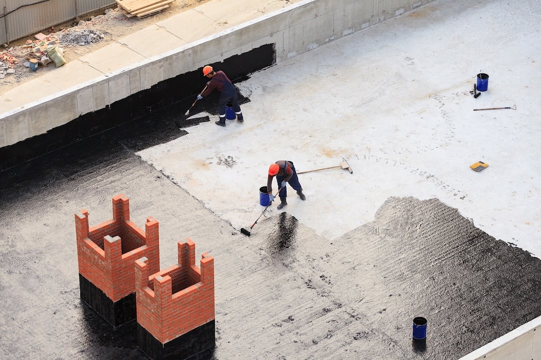 Two workers conduct waterproofing of flat roof with rollers.