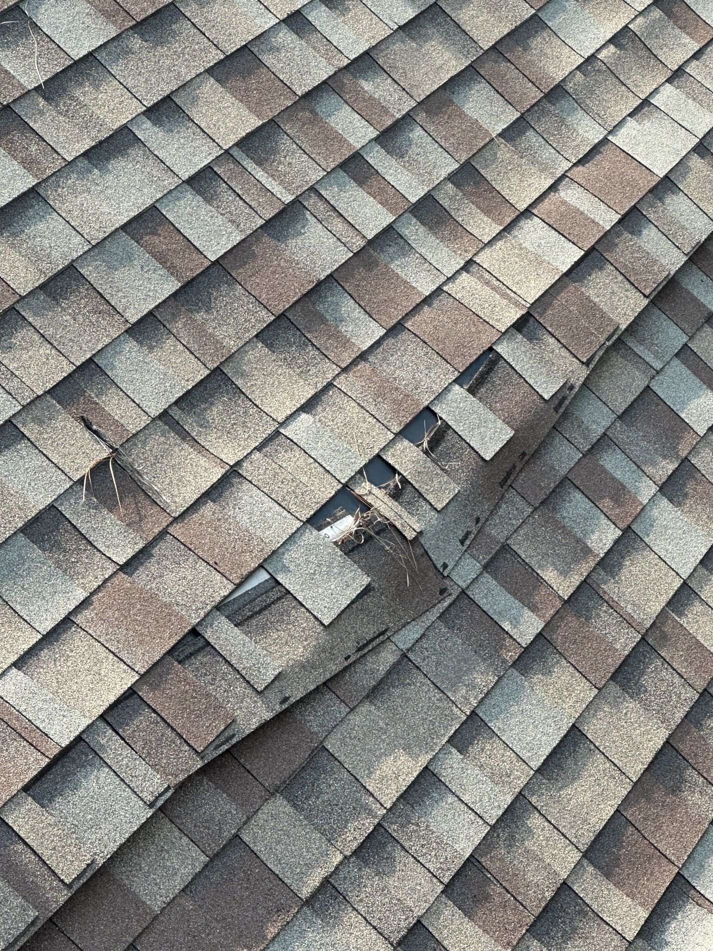 An asphalt shingled roof with a noticeable gap, exposing the structure underneath. Several shingles appear to be misaligned or missing, indicating potential damage or wear. There is some debris visible within the opening.