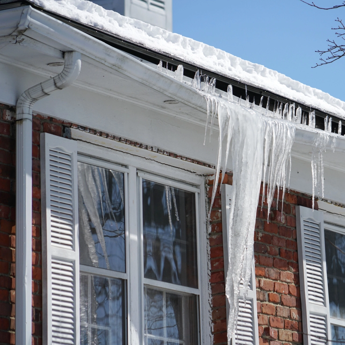 A brick house with white shutters and two windows is topped with snow. Several large icicles hang from the edge of the roof, sparkling in the sunlight against a clear blue sky. The scene evokes a cold winter day.