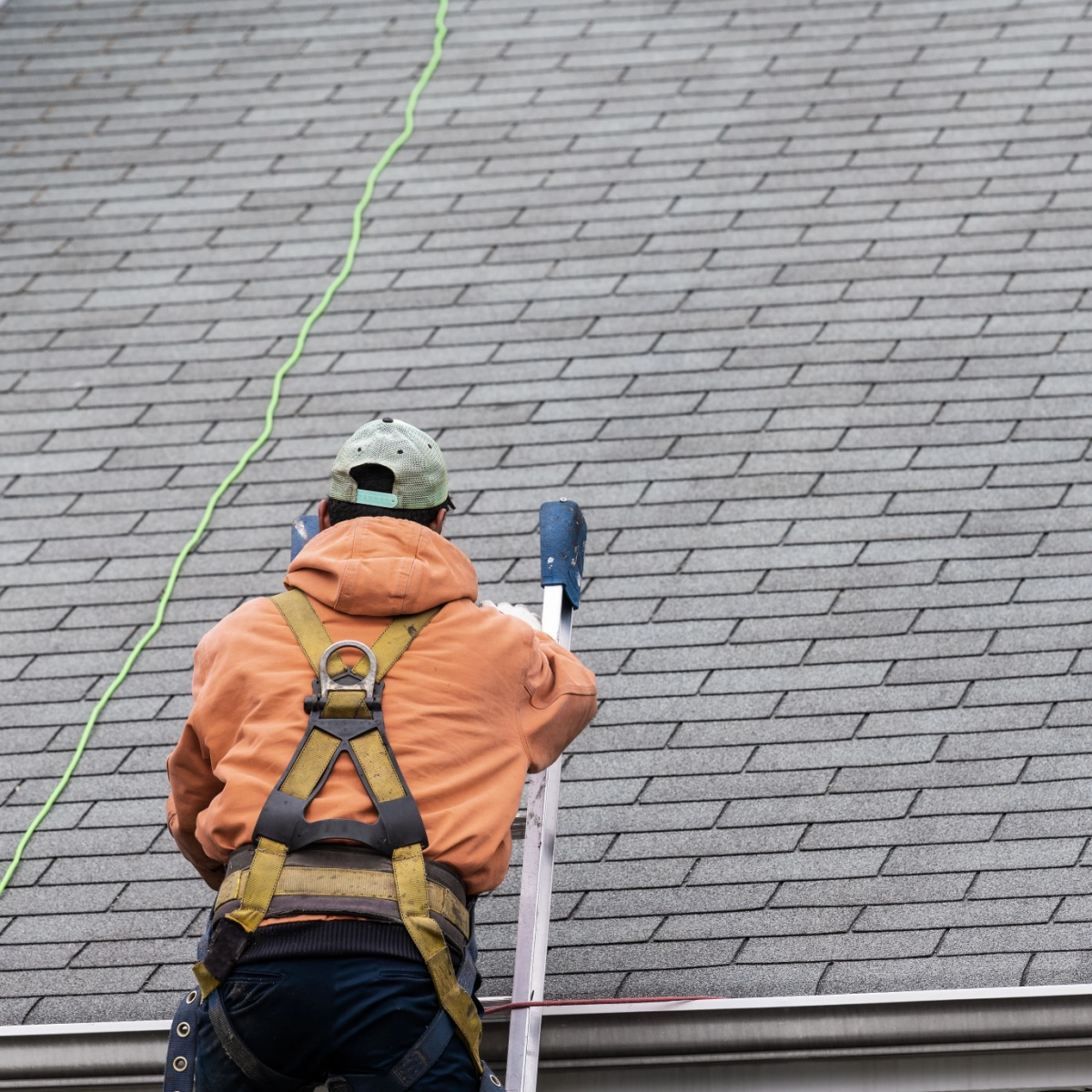 A person in an orange hoodie and safety harness climbs a ladder onto a shingled roof, with a green safety line attached to their harness, likely preparing for a Houston roof replacement.