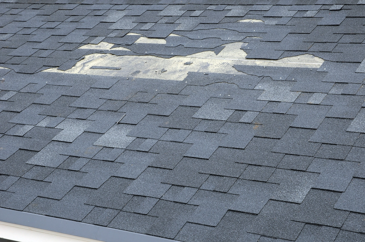 An asphalt shingle roof with noticeable damage, showing missing and worn shingles in a central section. The exposed underlayment is visible where the shingles have been stripped away. The surrounding shingles are intact but weathered, highlighting the need for professional Houston roofing services.