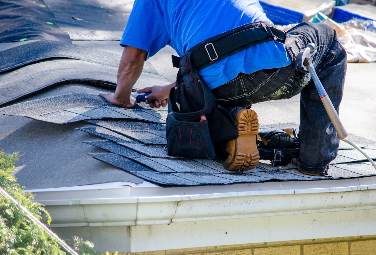 A person in a blue shirt and tool belt kneels on a rooftop, diligently installing shingles. They use a hammer and have various tools in their belt and scattered around them. The roof, partially covered with new shingles, reflects the quality work characteristic of Houston roofing professionals.