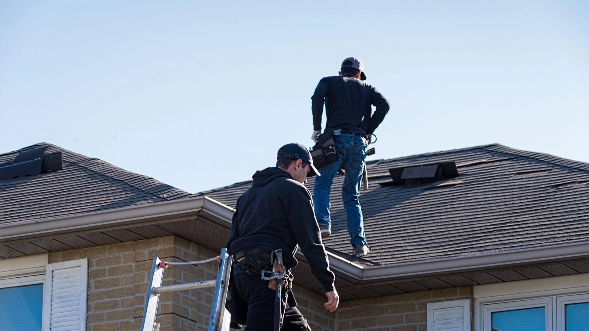 Two workers are conducting a Houston roof inspection. One is standing on the rooftop, looking around with hands on hips, while the other is climbing a ladder. Both are wearing safety harnesses and hats. The house is made of brick and has a shingled roof. The sky is clear.