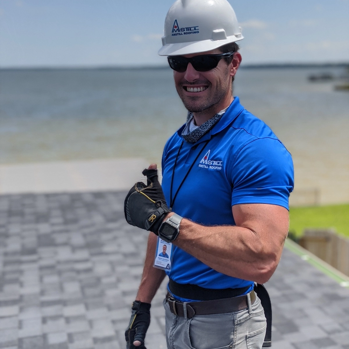 A person wearing a white hard hat, blue polo shirt, black gloves, and sunglasses smiles while standing on a roof under construction by Katy Roofing. They are giving a thumbs-up gesture, and the background shows a large body of water and a grassy area.