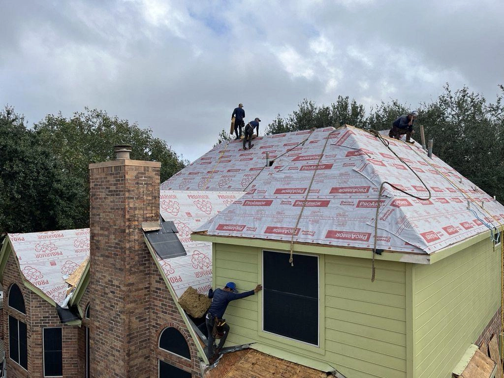 Roofers are diligently installing new shingles on a house with overlaid tar paper. The roof, part of a detailed Houston roof replacement, has a complex design with multiple slopes and dormers. The house features light green siding and a brick chimney. Workers, using tools and safety harnesses, are surrounded by trees under a cloudy sky.