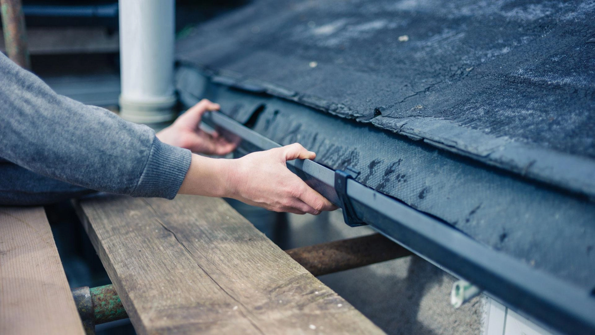 A person is installing a rain gutter under the edge of a weathered roof in Houston. They are holding the gutter in place with both hands while standing on a wooden platform. A white pipe can be seen in the background, characteristic of many Houston roofing projects.
