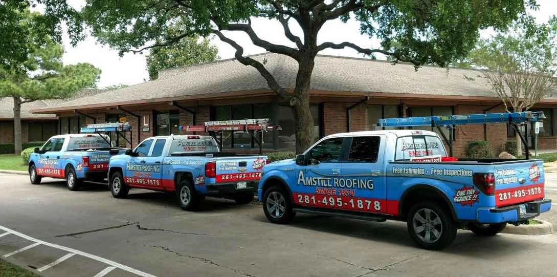 Three parked pickup trucks branded with "Amstill Roofing" logos and contact information. Each truck has a roof rack carrying ladders. The vehicles are parked side by side on a paved lot in front of a row of brick buildings, shaded by trees.
