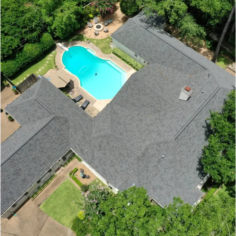 Aerial view of a residential property featuring a large, grey roofed house forming a rough V-shape around a spacious, curvy swimming pool, surrounded by a paved area with chairs and greenery. The property is bordered by trees and a driveway is visible on the left side.