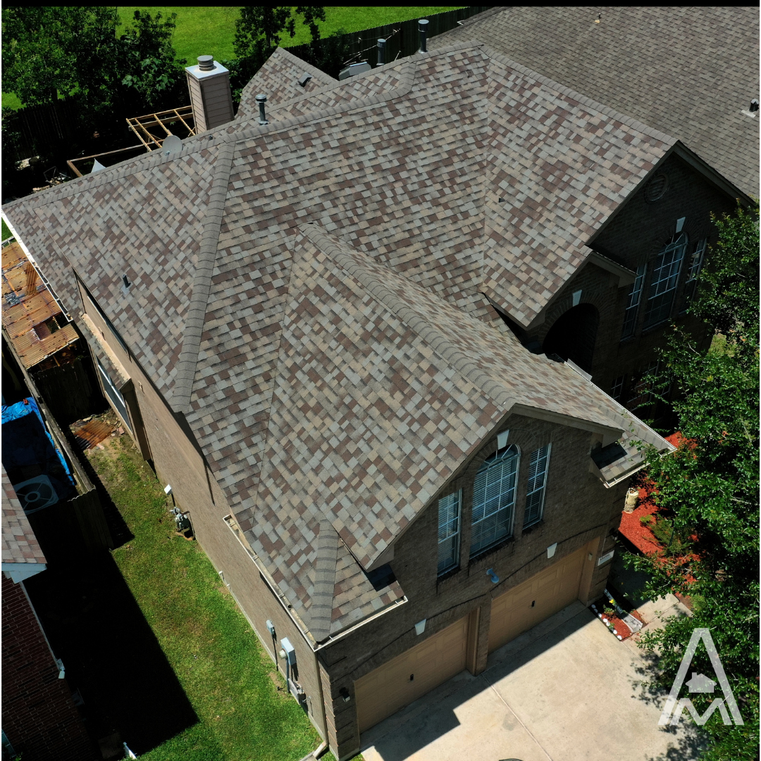 Aerial view of a large, two-story house with a steep, complex roof design covered in brown shingles. The property features a three-car garage and well-maintained surrounding lawn. "A" logo is visible in the bottom right corner.
