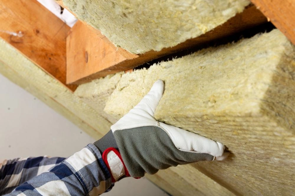 A person in a plaid shirt wearing gray and white gloves installs mineral wool or fiberglass insulation between wooden beams, possibly as a preventive measure against Tomball roof damage.