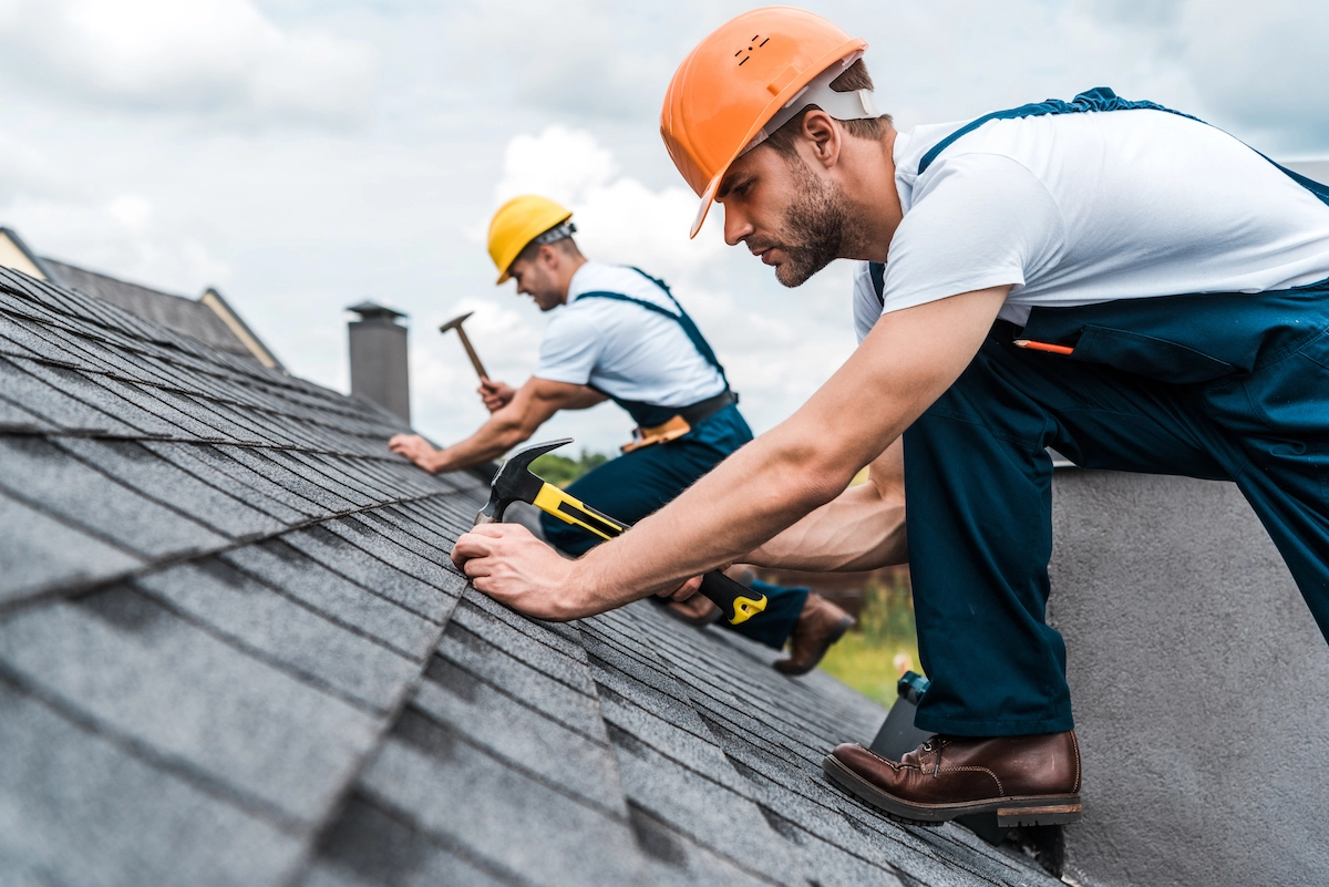 Two construction workers in overalls and safety helmets are working on a sloped roof in Houston. One is using a hammer while the other is positioning shingles, showcasing their expertise in roof repairs. The sky is partly cloudy in the background.