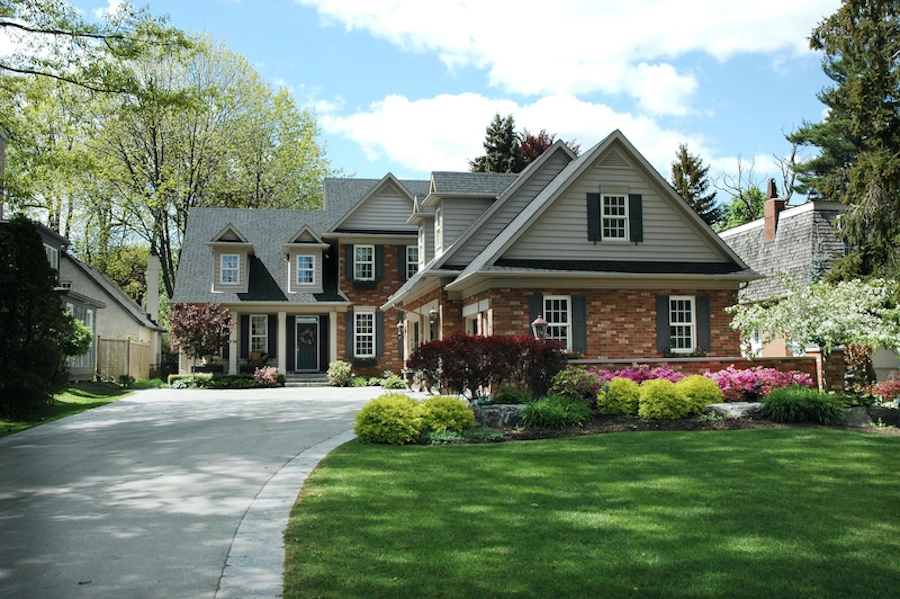 A two-story suburban house with a manicured front lawn and garden. The house features a mix of brick and siding exterior, multiple windows with shutters, and an attached garage. A curved driveway leads up to the front entrance, showcasing a property surrounded by green trees and perfect for Houston roof repair professionals to service.