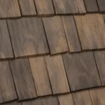 The image shows a close-up view of wooden shingles on a roof. The shingles have a natural brown color with visible grain and texture, creating a rustic and sturdy appearance. They are arranged in overlapping rows, providing a weathered and traditional look.