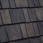 A close-up view of a grey wooden shingle roof with overlapping rectangular shingles, each displaying natural wood grain and texture. The shingles are uniformly arranged, creating a pattern on the roof surface.