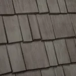 A close-up view of a roof with wooden shingles. The shingles are arranged in overlapping rows, each shingle having a slightly weathered appearance with natural grain textures visible. The overall color is a muted, earthy brown.