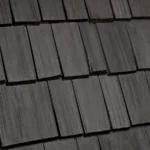 A close-up view of a dark wooden shingle roof displaying a uniform pattern of rectangular, weathered shingles. The texture and grain of the wood are visible, contributing to a rustic aesthetic.