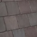 A close-up image of a textured slate roof, featuring uniform, overlapping rectangular slate tiles with a blend of dark and light gray hues. The tiles have a slightly uneven surface, giving a natural, rustic appearance.