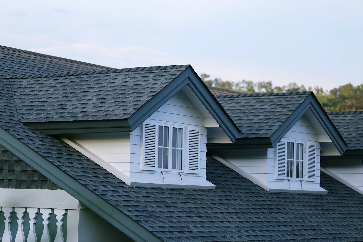 A close-up of a house roof by Rosenberg Roofing, featuring two dormer windows with white shutters, gray asphalt shingles, and white siding. A balcony with white railings is visible on the left. Trees and a clear sky are in the background.