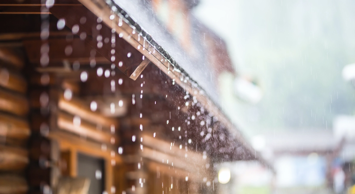 Raindrops falling from the eaves of a rustic wooden cabin evoke the craftsmanship of Spring TX roofing. The background includes blurred elements of the cabin's walls, with a hint of greenery and other buildings, suggesting a wet and misty day.