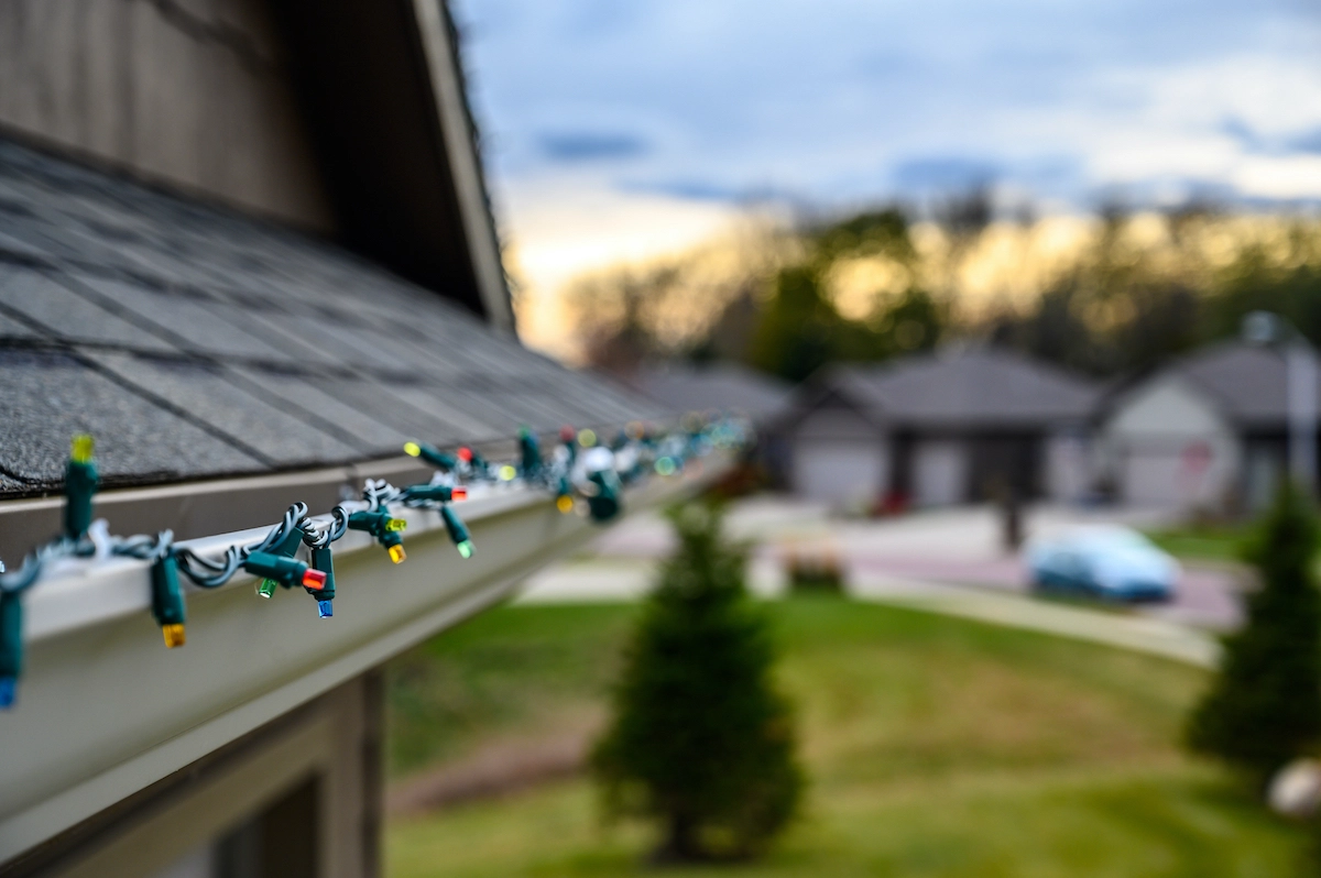 Close-up of a string of multicolored holiday lights hanging from the edge of a house roof. The background shows a blurred suburban neighborhood with homes, trees, and a parked car under a cloudy sky, subtly reminding one of the charm and needs for Houston roof repair in festive times.